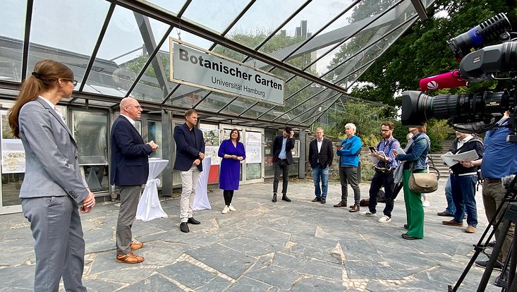 Pressekonferenz vor den Schaugewächshäusern am Dammtor. Eine Gruppe von Menschen im Halbkreis vor dem Bau aus Glas und Stahl
