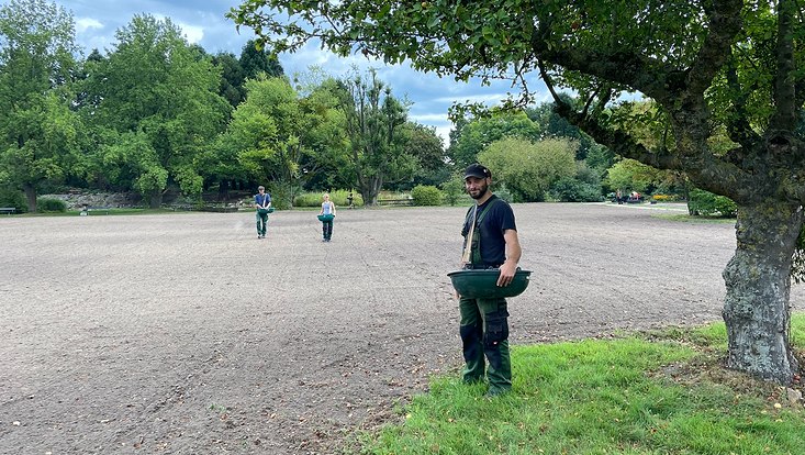 Blick auf eine große, umgepflügte Fläche, im Vordergrund ein Baum, im Hintergrud sääen zwei Personen Saat aus